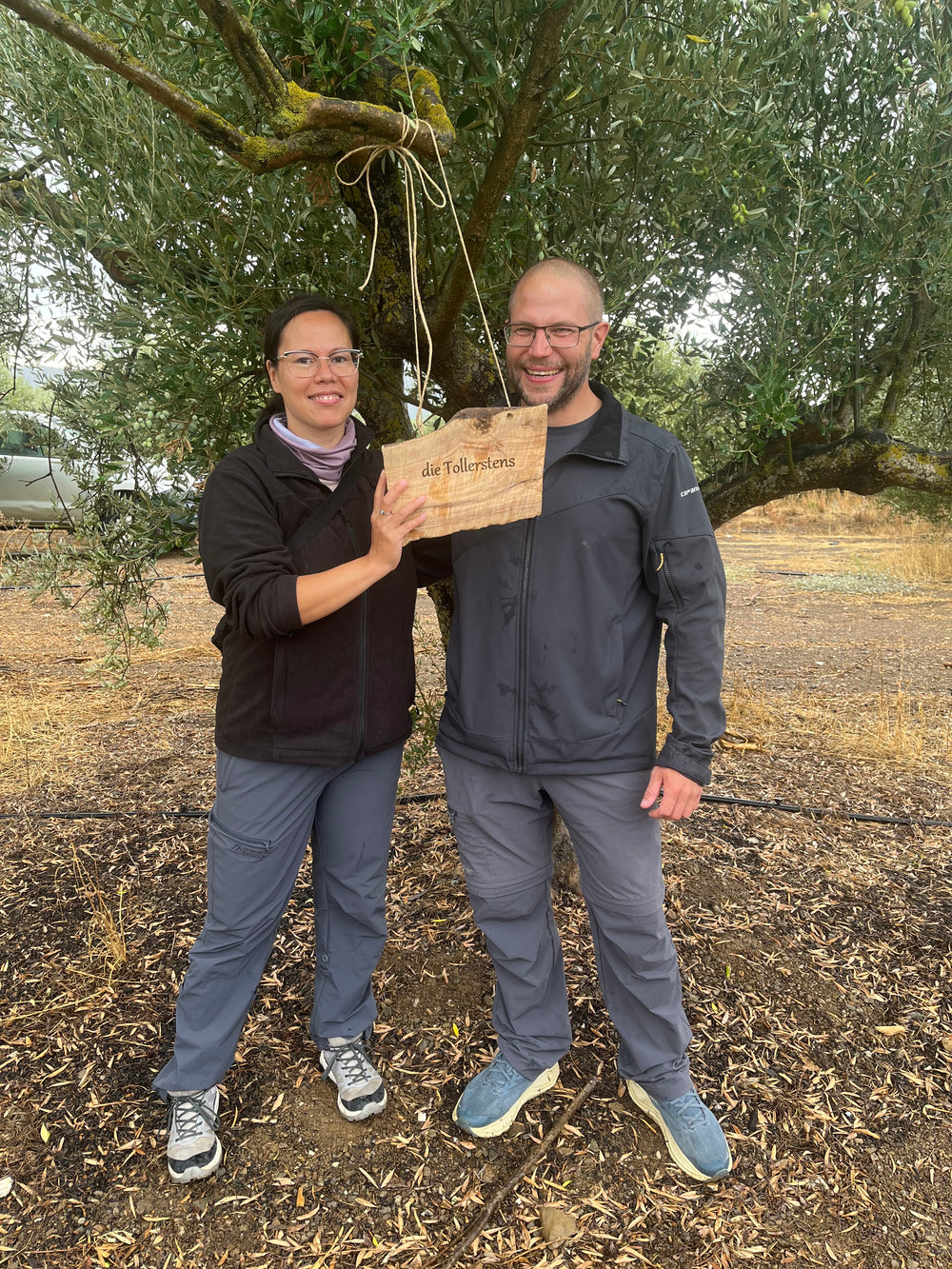 A couple from Germany in front of the Olive Tree they adopted in Crete by Olivine holding their personalised wooden plaque with their name on it